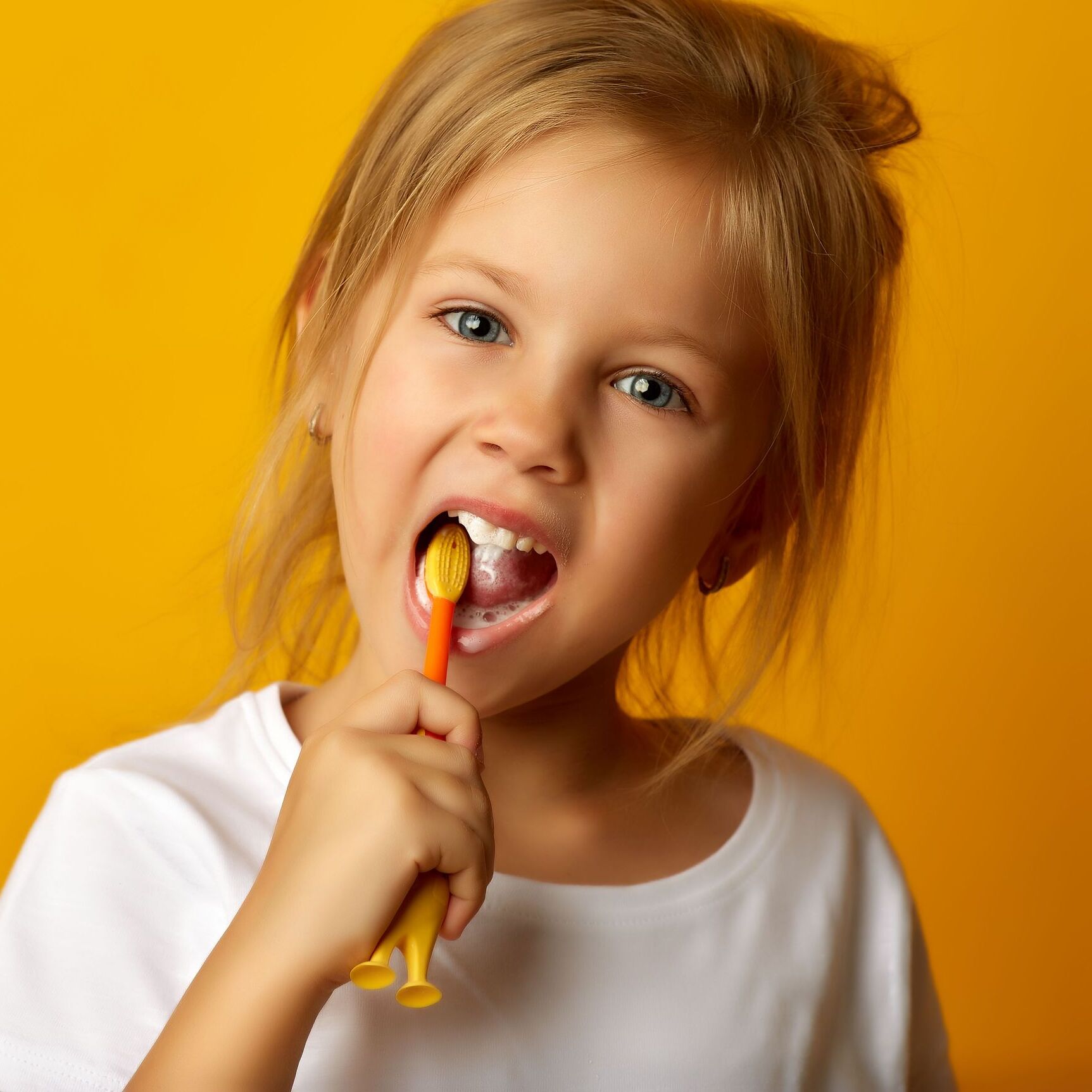 Junges blondes Mädchen putzt sich glücklich vor einem gelben Hintergrund die Milchzähne mit einer Kinder-Zahnbürste